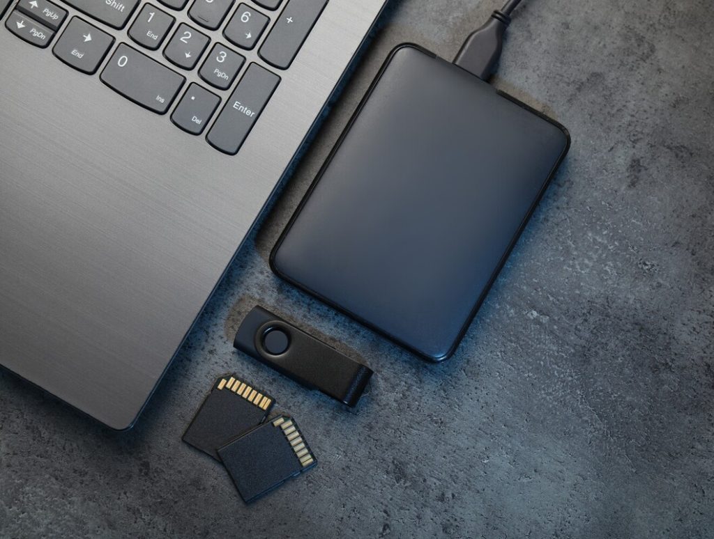 SD memory cards, USB flash drive, hard disk drive with cable and laptop on dark grey table, flat lay. 