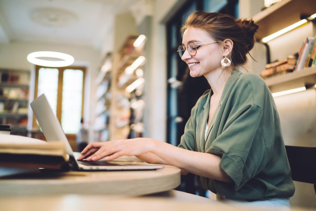 Cheerful young freelance woman typing on laptop in library